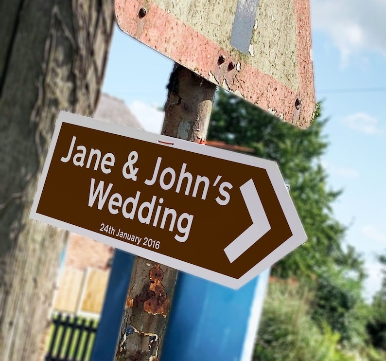 May include: A brown directional sign with white text that reads "Jane & John's Wedding" and "24th January 2016". The sign has a white arrow pointing to the right. The sign is mounted on a weathered metal pole.