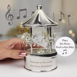 Peut inclure: Bo&icirc;te &agrave; musique en argent en forme de carrousel avec une base blanche. La base est grav&eacute;e du texte "Bienvenue au monde Ava Grace Nicholson". Le carrousel joue la chanson "Rock-a-bye Baby".