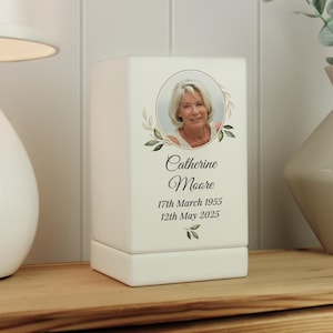 May include: A white memorial box featuring a photograph of a woman within a gold circular frame, adorned with a green leaf motif. The box displays the name "Catherine Moore" and the dates "17th March 1955" and "12th May 2025".