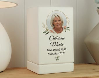 May include: A white memorial box with a photo of a woman in a gold frame, surrounded by a green leaf design. The text reads "Catherine Moore" and dates: "17th March 1955" and "12th May 2025".