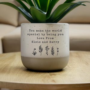 May include: A two-tone ceramic plant pot with a green plant. The top half is off-white with black text: "You make the world special by being you. Love From Elsie and Harry." The bottom half is light brown, with floral illustrations.