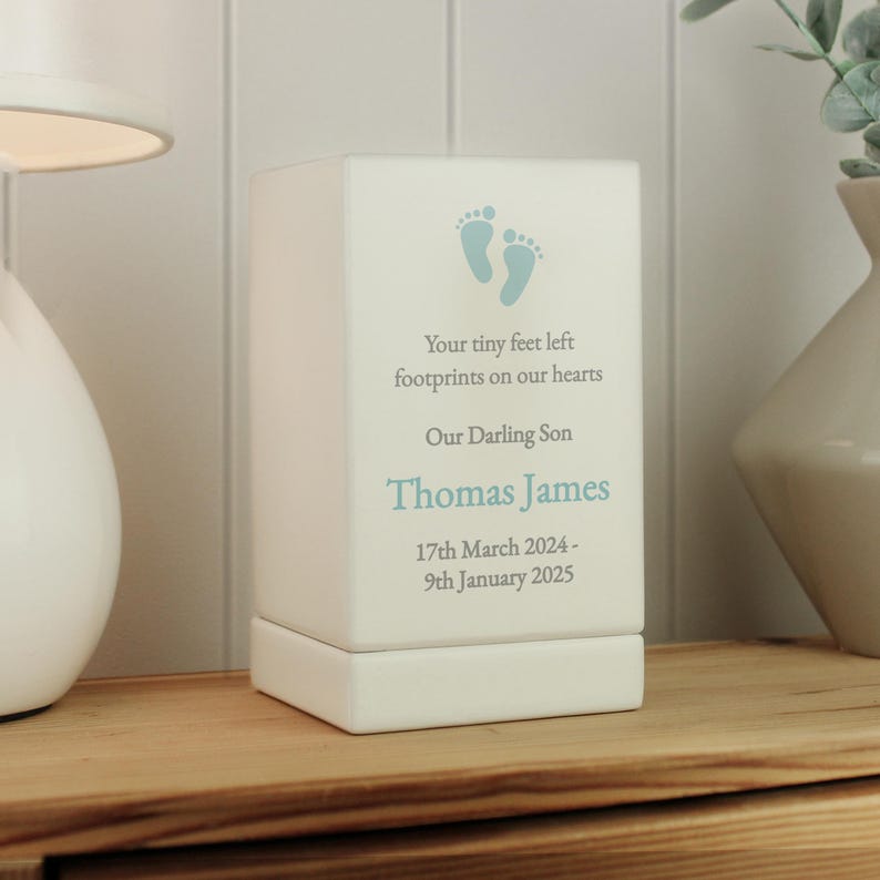 Może przedstawiać: Biała ceramiczna skrzynka na pamiątki z niebieskim wzorem odcisku stopy. Skrzynka zawiera tekst "Your tiny feet left footprints on our hearts Our Darling Son Thomas James 17th March 2024 - 9th January 2025"
