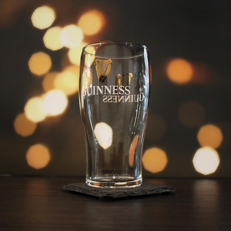 May include: A clear glass pint glass with the Guinness logo and harp printed on the side. The glass is sitting on a black slate coaster.