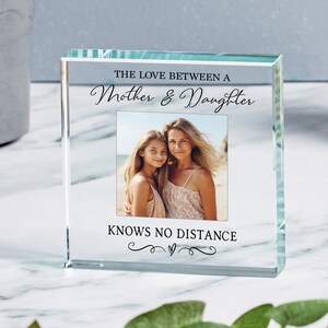 May include: A clear glass photo frame with a white background and black text that reads "The love between a mother & daughter knows no distance". The frame has a small photo of a mother and daughter in it.