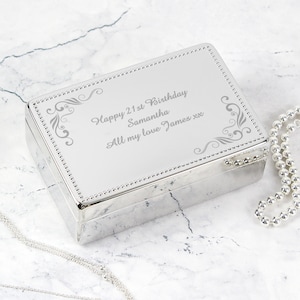May include: A silver rectangular jewellery box with a hinged lid. The lid is engraved with the text "Happy 21st Birthday Samantha All my love James xx" and decorative flourishes. A beaded border frames the lid. A silver necklace is draped nearby.