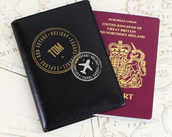 May include: A black passport holder with a gold embossed stamp that says "Bon Voyage, Holiday, Explore, Travel, Escape" and "Tom" in the center. The passport holder is lying on top of a map with a red British passport visible in the background.
