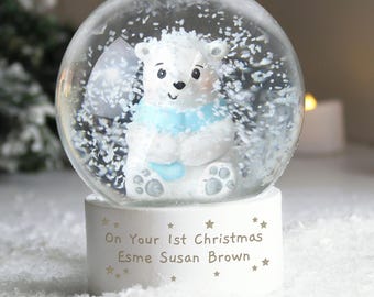 May include: A snow globe featuring a white polar bear wearing a light blue scarf. The bear sits inside a clear glass globe filled with white flakes. The white base has the text "On Your 1st Christmas Esme Susan Brown" with star accents.