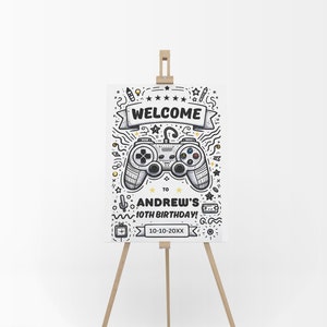 Puede incluir: Un cartel de bienvenida blanco con una ilustración en blanco y negro de un mando de videojuego. El cartel dice "¡Bienvenido al 10º cumpleaños de Andrew! 10-10-20XX". El cartel está en un caballete de madera.