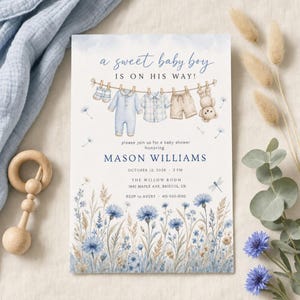 May include: A baby shower invitation card featuring the text A sweet baby boy is on his way, with illustrations of baby clothes on a clothesline, including a onesie, shirt, shorts, and teddy bear, above blue wildflowers and event details.