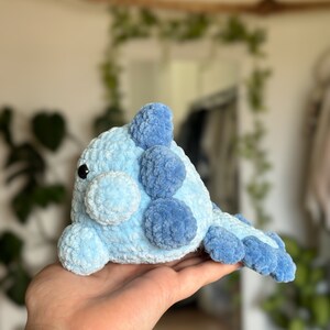 May include: A crochet plush toy shaped like a creature with a rounded body, featuring a light blue main colour and darker blue accents on the fins and tail. The toy has a soft, fuzzy texture, a small snout, and a single visible black plastic eye.