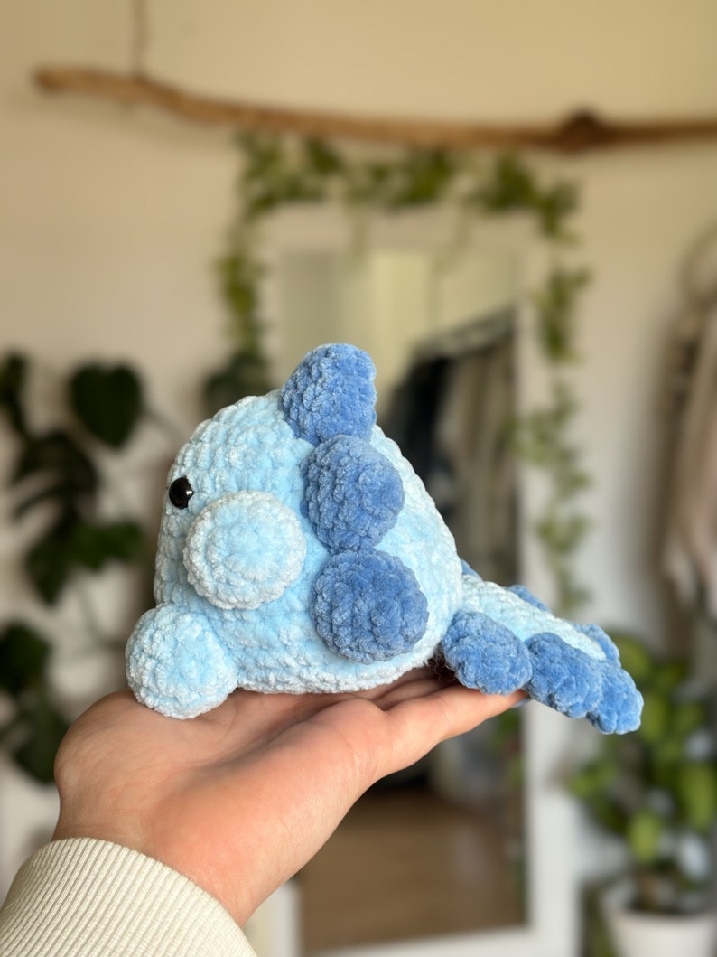 May include: A crochet plush toy shaped like a creature with a rounded body, featuring a light blue main colour and darker blue accents on the fins and tail. The toy has a soft, fuzzy texture, a small snout, and a single visible black plastic eye.