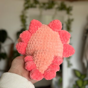 May include: A plush axolotl toy crafted from soft, fuzzy yarn in shades of light peach and vibrant pink. The creature features a rounded body with a central peach-coloured section and bright pink external gills extending from the sides and top.