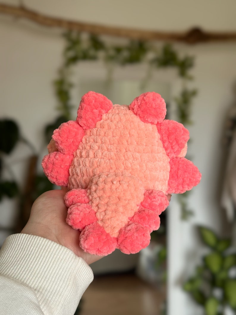 May include: A plush axolotl toy crafted from soft, fuzzy yarn in shades of light peach and vibrant pink. The creature features a rounded body with a central peach-coloured section and bright pink external gills extending from the sides and top.