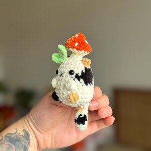 以下が含まれることがあります： 白地に黒と茶色の斑点がある牛の形をした、手編みの小さなかぎ針編みのぬいぐるみ。頭には鮮やかなオレンジ色のキノコの帽子をかぶっており、2つの小さな黒い目、明るい緑色の葉のアクセント、そして垂れ下がった尻尾が特徴です。