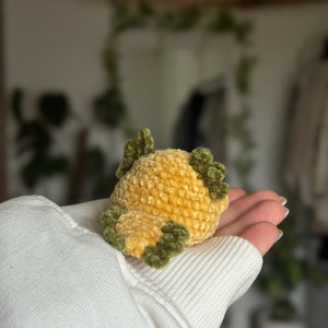 Peut inclure: Un petit jouet en peluche crochet&eacute; &agrave; la main, de forme ronde, pr&eacute;sentant un corps textur&eacute; jaune dor&eacute; avec des accents vert olive fonc&eacute; sur le dessus et les c&ocirc;t&eacute;s. Le fil doux et duveteux cr&eacute;e un motif de points dense et uniforme sur l'objet.
