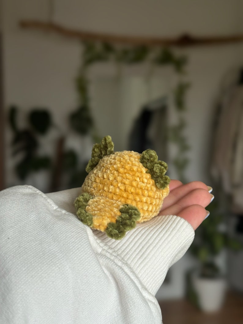 Peut inclure: Un petit jouet en peluche crochet&eacute; &agrave; la main, de forme ronde, pr&eacute;sentant un corps textur&eacute; jaune dor&eacute; avec des accents vert olive fonc&eacute; sur le dessus et les c&ocirc;t&eacute;s. Le fil doux et duveteux cr&eacute;e un motif de points dense et uniforme sur l'objet.