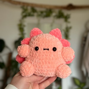 May include: A plush toy made of soft, light pink yarn with a rounded body, small limbs, and a row of darker pink, petal-like protrusions along the back. It features two small, round, black plastic eyes and a simple, straight black stitched mouth.