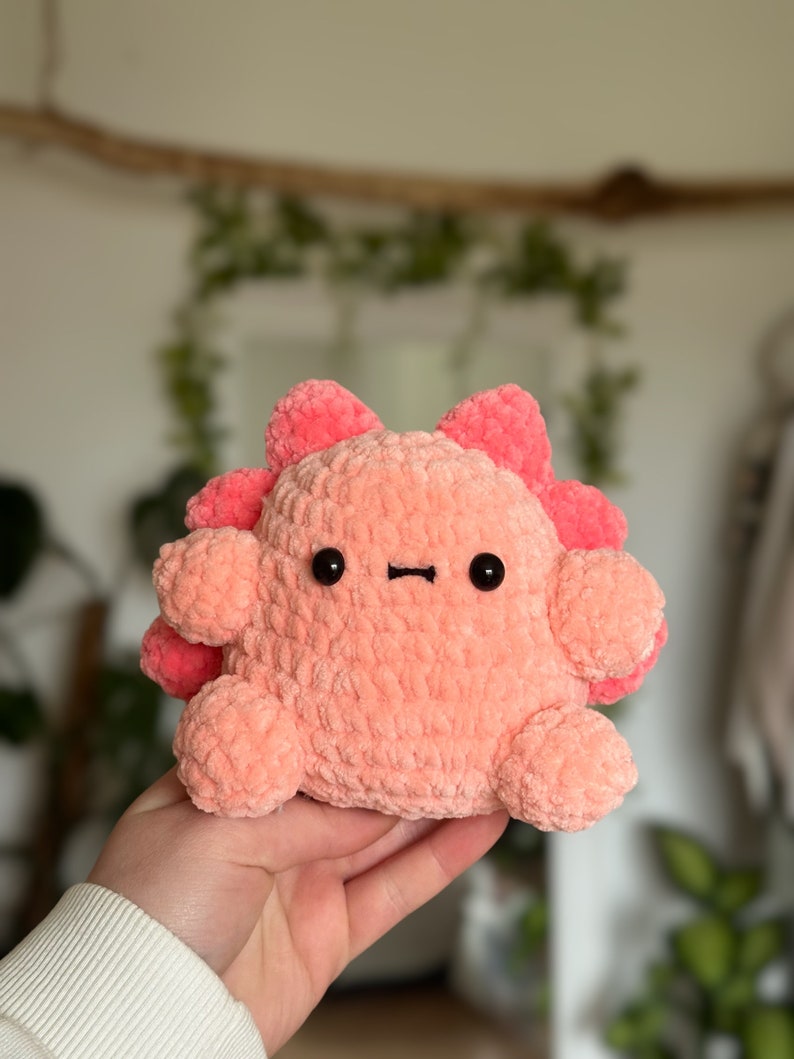 May include: A plush toy made of soft, light pink yarn with a rounded body, small limbs, and a row of darker pink, petal-like protrusions along the back. It features two small, round, black plastic eyes and a simple, straight black stitched mouth.