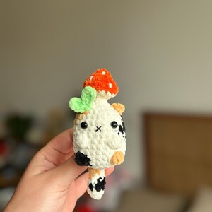 以下が含まれることがあります： 黒、白、オレンジの斑点がある三毛猫の形をした、手作りの小さなかぎ針編みのぬいぐるみ。猫には黒いビーズの目、刺繍された口があり、頭には白い斑点のある小さなオレンジ色のキノコと明るい緑色の葉が付いています。