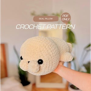 Puede incluir: Un cojín de foca tejido a ganchillo de color crema con grandes ojos redondos de plástico negro. El texto en la imagen dice: SEAL PILLOW, PDF ONLY, CROCHET PATTERN. La foca tiene una cabeza redondeada, un hocico pequeño y aletas visibles.