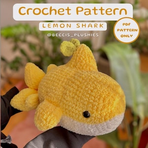 May include: A crocheted lemon shark plush toy in yellow and white. The toy has a small, green fin on top of its head. The image includes the text "Crochet Pattern Lemon Shark @beccis_plushies PDF Pattern Only".