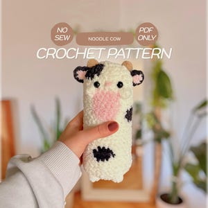 May include: A white, black, and pink crochet cow plushie with small brown horns and black eyes. The text NO SEW, PDF ONLY, NOODLE COW, and CROCHET PATTERN is displayed above the plushie. The cow has a cylindrical body with black spots.