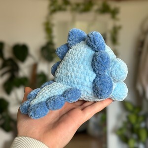 May include: A small, plush, crocheted dinosaur toy crafted from soft, light blue yarn. The creature features a row of darker blue, rounded, textured spikes running along its back and down its tail, creating a distinct, tactile, and soft appearance.