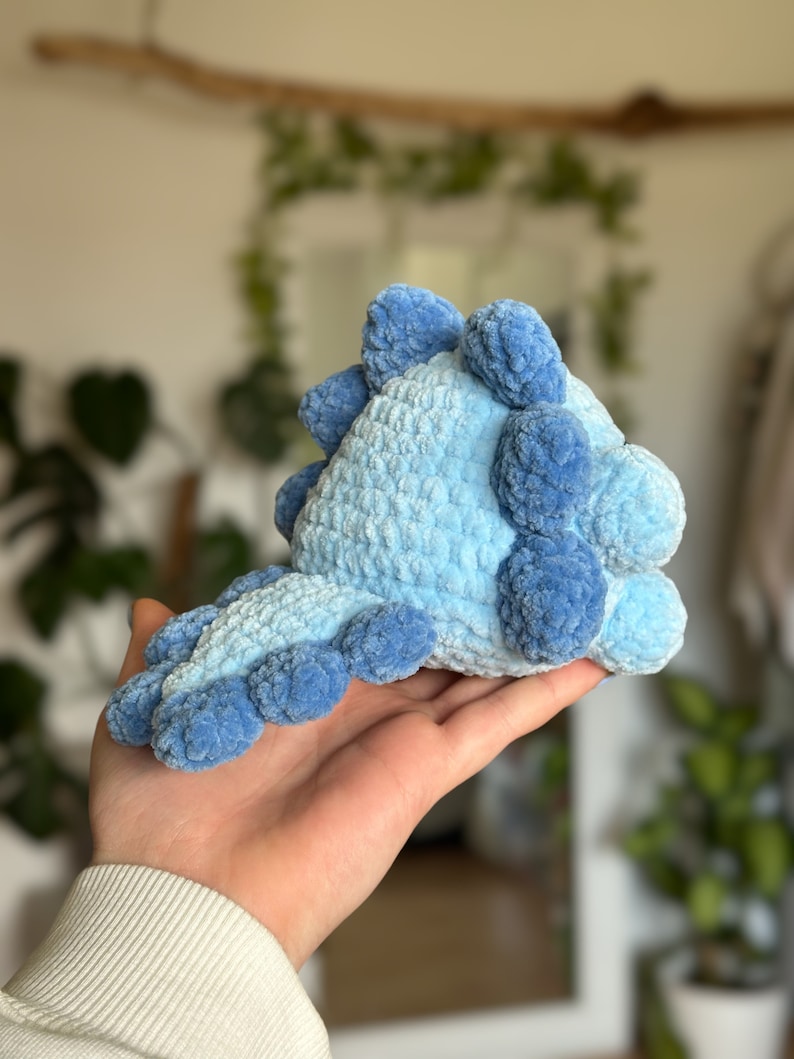 May include: A small, plush, crocheted dinosaur toy crafted from soft, light blue yarn. The creature features a row of darker blue, rounded, textured spikes running along its back and down its tail, creating a distinct, tactile, and soft appearance.