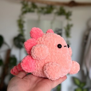 May include: A plush toy shaped like a dinosaur, crafted from soft, fuzzy light pink yarn. It features darker pink rounded spikes along its back and tail, two small black circular eyes, and a rounded body with short, stubby limbs.
