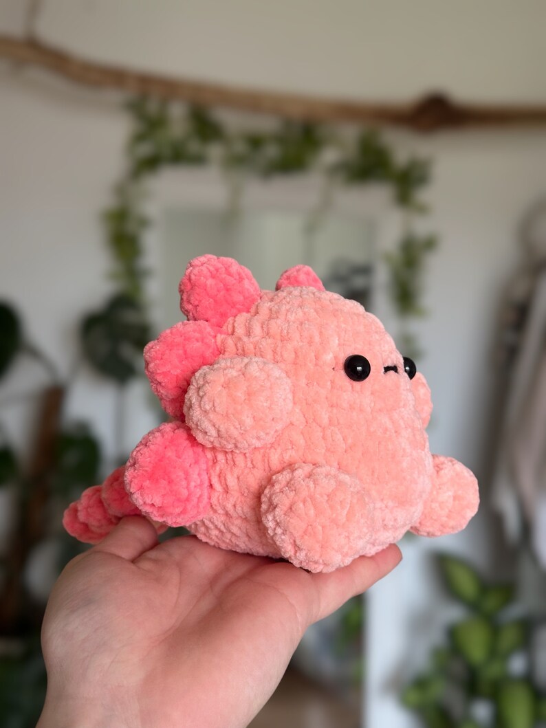 May include: A plush toy shaped like a dinosaur, crafted from soft, fuzzy light pink yarn. It features darker pink rounded spikes along its back and tail, two small black circular eyes, and a rounded body with short, stubby limbs.