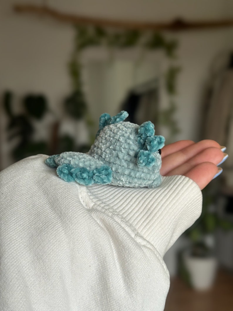 Peut inclure: Un petit axolotl en crochet fait main, r&eacute;alis&eacute; avec un fil de velours bleu clair doux. La cr&eacute;ature pr&eacute;sente une t&ecirc;te arrondie, une queue plate et des branchies externes bleu sarcelle plus fonc&eacute;es sur les c&ocirc;t&eacute;s, avec un &oelig;il noir visible.