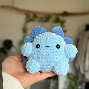 May include: A small, round, crocheted plush toy in shades of light and medium blue. The creature features a textured, soft yarn surface, two small black plastic eyes, a simple horizontal stitched mouth, and rounded appendages with a scalloped back.