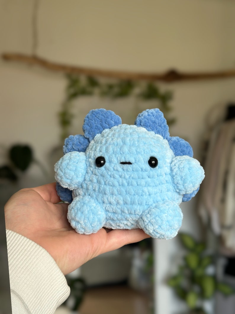 May include: A small, round, crocheted plush toy in shades of light and medium blue. The creature features a textured, soft yarn surface, two small black plastic eyes, a simple horizontal stitched mouth, and rounded appendages with a scalloped back.