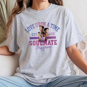 Könnte beinhalten: Ein graumeliertes T-Shirt mit Rundhalsausschnitt und einem Grafikdruck mit dem Text LOVE UPON A TIME, EST 2026, SOULMATE Program sowie einer zentralen Collage aus vier Personen in verschiedenen Posen in Violett-, Blau- und Rosatönen.