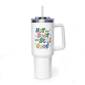 May include: White insulated travel mug with a handle and a straw. The mug has the text "NOT the BEST but STILL GOOD" in colourful letters with star and heart accents. The lid is blue.