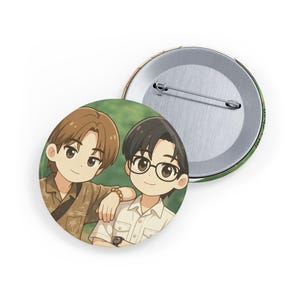 Könnte beinhalten: Ein runder Ansteckbutton mit einer Illustration von zwei Zeichentrickfiguren. Eine Figur hat braune Haare und ein beiges Hemd, die andere hat schwarze Haare und eine Brille und trägt ein weißes Hemd. Die Rückseite des Buttons ist aus Metall mit einer Sicherheitsnadel.