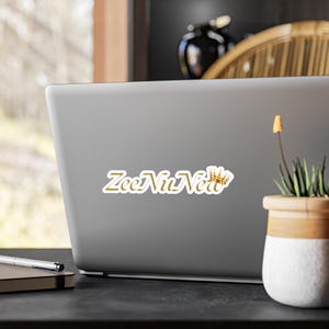 May include: A sticker on a laptop with the text "ZeeNuNerd" in gold script, accompanied by a small gold crown graphic. The laptop is gray and sits on a dark surface.