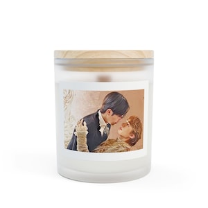 May include: A white glass candle with a wooden lid. The candle features a label with a romantic image of two people. The image shows two people in formal attire, looking at each other. The candle is a home decor item.