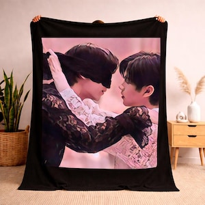 May include: Black blanket featuring a digitally painted image of two people embracing. One person is wearing a black blindfold. The image has a soft, pink and white background. The blanket is held up by unseen hands.