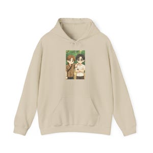 Peut inclure: Un sweat à capuche beige avec une poche avant. Le sweat-shirt présente un graphisme rectangulaire de deux personnages de style anime. Un personnage porte une chemise à motifs, et l'autre porte une chemise blanche et des lunettes.