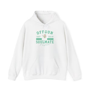 Puede incluir: Sudadera con capucha blanca con bolsillo delantero y cordón. La sudadera presenta el texto "OFFGUN" encima de "SOULMATE Program" en verde, con "EST 2019" y un pequeño gráfico en medio. Una prenda casual y cómoda.