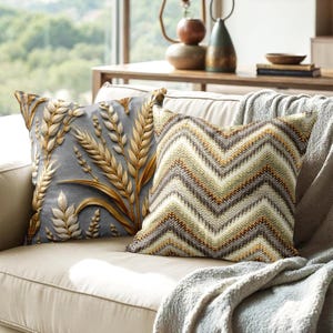 Könnte beinhalten: Zwei dekorative Kissen auf einem beigen Sofa. Ein Kissen hat einen grauen Hintergrund mit einem goldenen Weizenmuster. Das andere Kissen hat ein Chevron-Muster in Braun-, Beige- und Weißtönen.