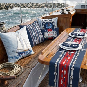 Op de afbeelding: Nautische decoratie op een bootdek. Inclusief kussens met zeilboot- en touwontwerpen, een tafelloper met vis- en touwdetails en servies. Het kleurenschema is marineblauw, wit en rood.