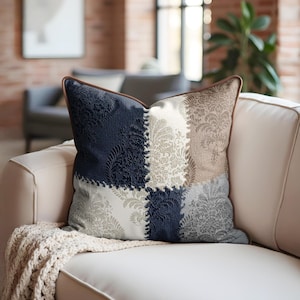 May include: Square cushion with a patchwork design. The cushion features four distinct sections in navy blue, beige, grey, and white, each with a damask pattern. The cushion has a brown trim and is placed on a cream-coloured sofa.