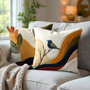 Peut inclure: Deux coussins décoratifs avec des motifs brodés. Un coussin représente un oiseau sur une branche avec un fond abstrait coloré. L'autre coussin a un motif feuillu avec un fond jaune et brun.