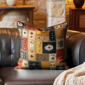 May include: Square decorative pillow with a patchwork design in shades of yellow, red, blue, and brown. The pillow is placed on a dark brown leather couch with a cream-colored blanket draped over the arm. The pillow has a brown border.