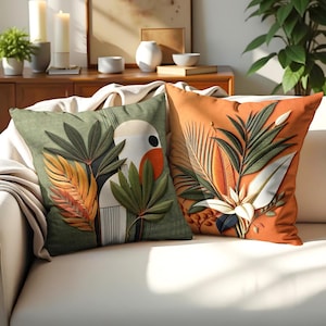 Op de afbeelding: Twee decoratieve kussens met een tropisch bladontwerp. Een kussen is groen met oranje en witte accenten, de andere is oranje met witte en groene accenten.