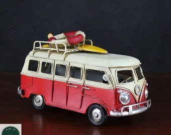 May include: A red and cream-colored vintage van model with a surfboard on top. A small figure in red and white clothing is lying on the surfboard. The van has a luggage rack and is on a wooden surface.