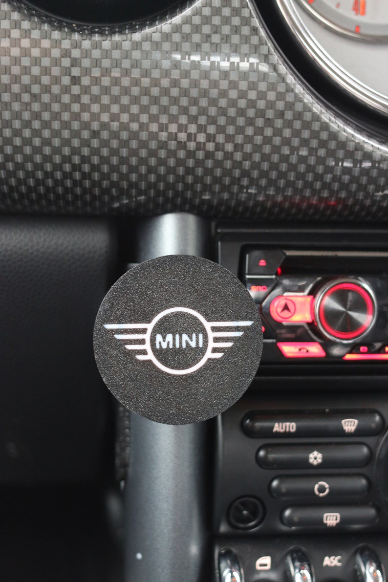 Peut inclure: Un bouton rond noir avec le mot "MINI" en lettres blanches et un design d'aile stylis&eacute; de chaque c&ocirc;t&eacute; du mot. Le bouton est fix&eacute; &agrave; un tableau de bord de voiture noir et argent.