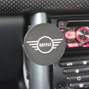 Peut inclure: Un accessoire de voiture rond noir avec le mot "MINI" en lettres blanches et un logo ail&eacute; argent&eacute;.